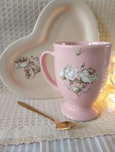 Taza chocolatera, color rosa. Incluye una cuchara de diseño, color dorada, y un plato de cerámica en forma de corazón.