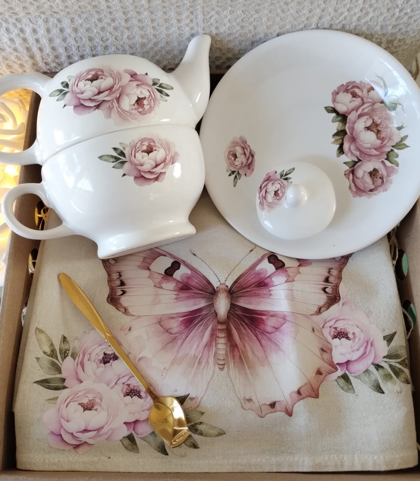 Box con set de té, con tazon, tetera, plato redondo, de cerámica con estampado de flores. Incluye cucharita de diseño, dorada, y mantel individual de lienzo de algodon, estampado con flores y mariposas